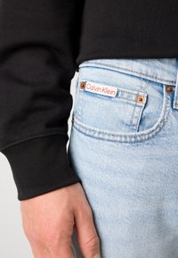 Käsi lepää vaaleansinisten farkkujen etutaskun lähellä, näkyvissä Calvin Klein -merkki ja musta pitkähihaisen paidan hiha.