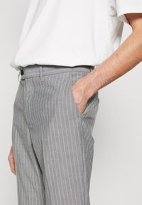 Šedé pruhované kalhoty slim fit s boční kapsou a zapínáním na knoflík. Materiál má hladkou texturu a jemný lesk.