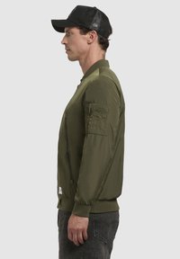 Maslinasto zeleni bomber jakna sa prednjim patent zatvaračem, rebrastim manžetama i bočnom džepom. Karakterizira je glatka, lagana tkanina i prilagođen dizajn.