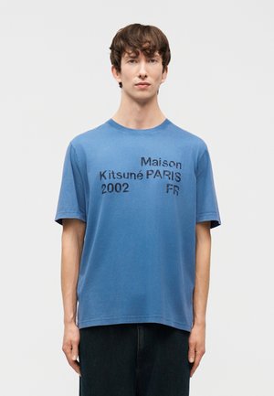 Jeune homme portant un t-shirt bleu Maison Kitsuné avec le texte noir "Maison Kitsuné PARIS 2002 FR" debout devant un fond uni.