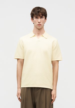 Νέος άνδρας με κοντά καστανά μαλλιά, φορώντας ένα μπεζ κοντομάνικο πόλο πουκάμισο με υφή και φαρδιά καφέ παντελόνια, στέκεται μπροστά σε απλό φόντο.