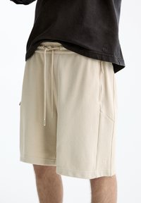 Beige drawstring shorts with side zipper pockets worn with a black casual shirt, showing mid-thigh length and relaxed fit.