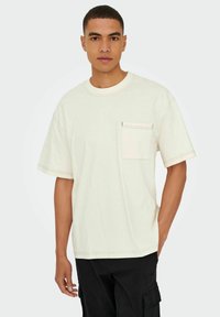Kortærmet, beige bomulds t-shirt med en brystlomme, kontrastsyninger og rund halsudskæring. Løs pasform og glat tekstur.