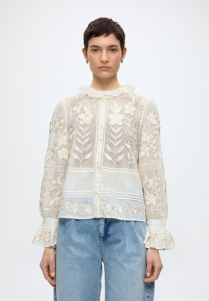 Mujer con blusa blanca de encaje con bordado floral, cuello y puños con volantes, combinada con jeans azul claro, de pie sobre fondo blanco.