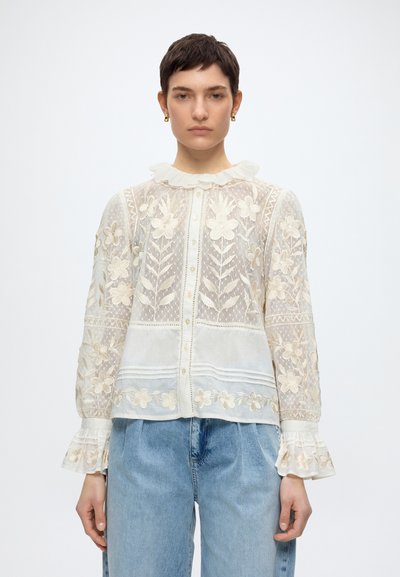 Femme portant un chemisier en dentelle blanche avec broderie florale, col et poignets à volants, associé à un jean bleu clair, debout devant un fond blanc.