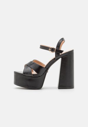 Black platform sandals featuring a glossy faux leather finish, thick block heels, and crisscross straps with a buckle closure.