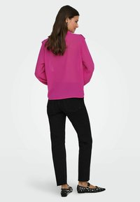 Fuchsia Bluse mit strukturierten Pünktchen, langen Ärmeln und lockerer Passform, kombiniert mit schwarzen Jeans und mit Nieten besetzten schwarzen Ballerinas.