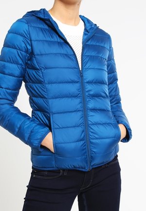 Person wearing a bright blue quilted hooded jacket with zipped front and hands in pockets, paired with dark jeans and a white shirt.