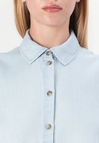 Ανοιχτό μπλε τζιν πουκάμισο με κλασικό γιακά, διαθέτει πέντε μεταλλικά κουμπιά στη μπροστινή πλευρά και είναι φτιαγμένο από μαλακό, υφαντό ύφασμα.