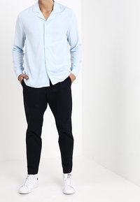 Chemise bleu clair à col cranté, associée à un pantalon noir ajusté et des baskets blanches, sur fond neutre.