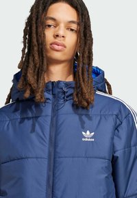 Jūrinė pūkinė striukė su užtrauktuku, kapišonu ir balta Adidas logotipu. Yra siuvinėta struktūra ir kontrastinga mėlyna pamušalė. Balti dryžiai ant pečių.