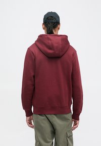 Sweatshirt burgundy com um bolso canguru na frente, punhos canelados e capuz com cordão, emparelhado com calças cargo em oliva.
