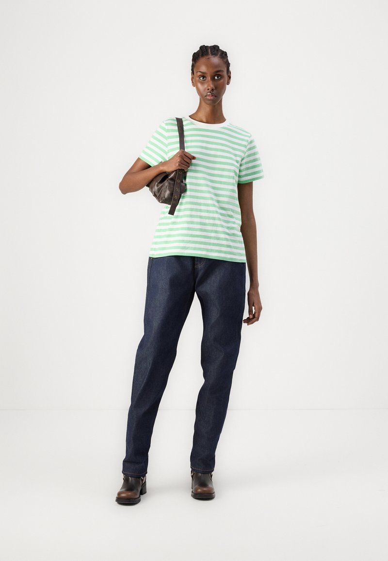 Camiseta de algodón a rayas verde y blanca, de corte relajado, combinada con jeans de mezclilla azul oscuro. La modelo sostiene un pequeño bolso marrón.