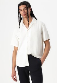 Camisa de manga corta en tela texturizada color blanco roto con un ajuste relajado, que presenta un cuello y cierre de botones, combinada con pantalones oscuros.