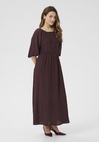 Robe maxi en bourgogne foncé, avec un corsage smocké, des manches courtes bouffantes et une jupe plissée et fluide. Associée à des escarpins rouges à bout pointu.