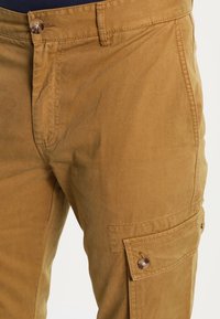 Khaki kleurige cargobroek met een knoopsluiting in de taille en een zijflapzak, gedragen met een donker shirt opgestoken.