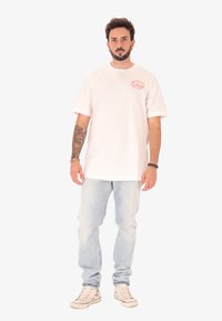 T-shirt en coton blanc à manches courtes, avec un petit logo rond rose sur la partie gauche de la poitrine, porté avec un jean bleu clair et des baskets beiges.