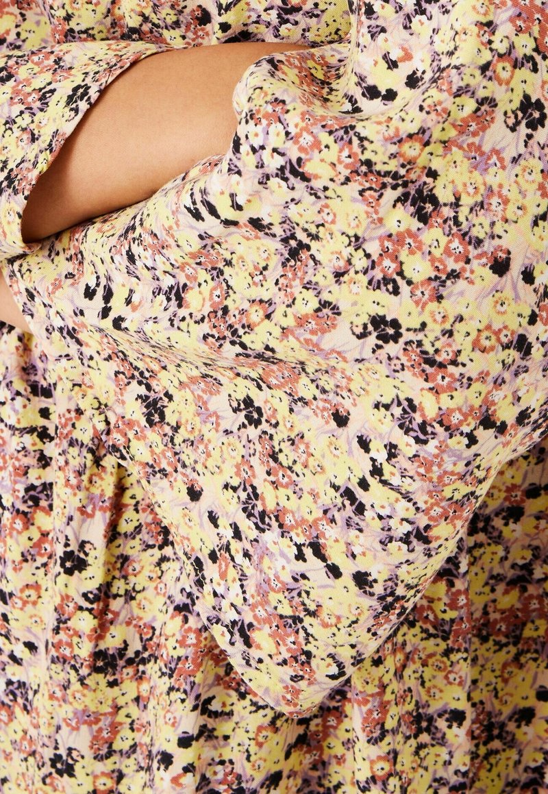 Květinové šaty z žluté látky se vzorem růžových, oranžových a černých květin. Volný střih s širokými volánovými rukávy.