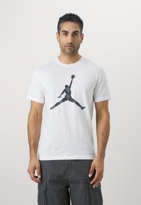 Hvid bomulds T-shirt med en sort silhuetgrafik af en springende figur, der holder en basketball. Kortærmet og med rund hals.