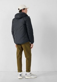 Uomo che indossa un giubbotto imbottito nero con cappuccio, pantaloni verde oliva arrotolati, sneakers bianche e un cappello bianco, in piedi contro un muro bianco semplice.