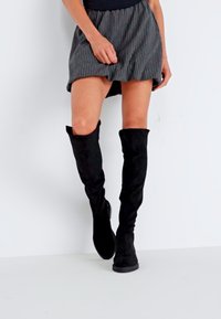 Cizme negre peste genunchi, dintr-un material suede moale, cu o formă ajustată și talpă plată, asortate cu o fustă rayată.