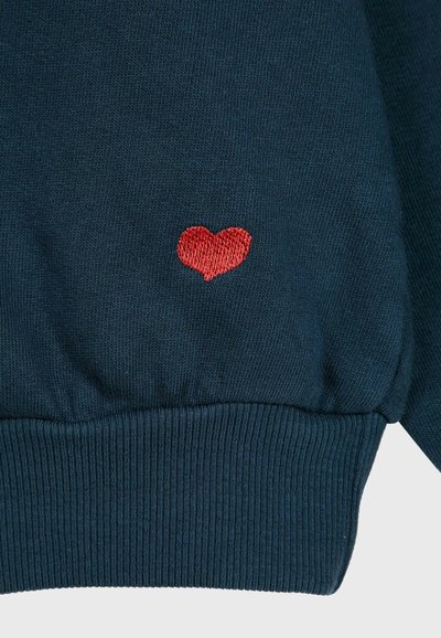 Dunkelblau gestrickter Stoff mit geripptem Saum und kleinem roten gestickten Herzen, das zentriert über dem Saum platziert ist.