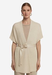 Beige stickad omslagscardigan med korta ärmar, bälte i midjan, öppen front och subtil lyster. Matchas med en ljus topp.