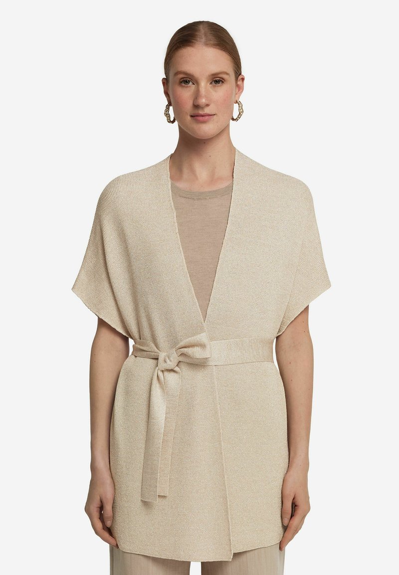 Beige stickad omslagscardigan med korta ärmar, bälte i midjan, öppen front och subtil lyster. Matchas med en ljus topp.