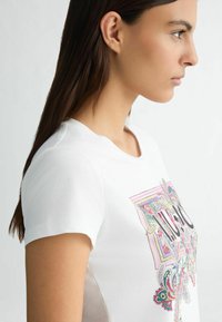 T-shirt bianco in cotone con un design grafico colorato sul davanti, scollo rotondo e maniche corte, mostrato in vista laterale.
