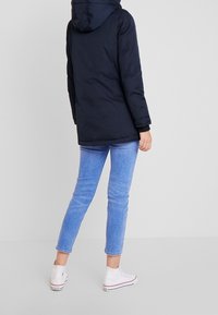 Marininspirerad vadderad kappa med huva, som har en slät textur och långa ärmar, kombinerad med ljusblå jeans och vita högskor.