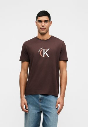 Bruine katoenen t-shirt met een wit en koperen "CK" logo, klassiek ronde halsontwerp en korte mouwen, gecombineerd met lichtblauwe jeans.