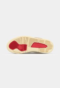 Semelle de sneaker avec une base en caoutchouc beige, présentant un accent rouge et des motifs de grip texturés, mettant en avant des éléments de design d'adhérence.