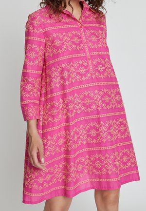 Vrouw die een roze jurk draagt met beige bloemmotieven, een kraag, driekwart mouwen en knopen aan de voorkant, staand tegen een effen achtergrond.