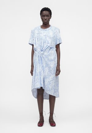 Femme portant une robe bleu clair brodée de fleurs, taille cintrée et ourlet asymétrique, associée à des chaussures plates rouges à lacets.