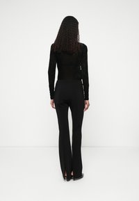 Črna dolga majica z teksturiranim finišem v kombinaciji z visokimi črnimi hlačami z bočnimi razporki, prikazano zadaj.