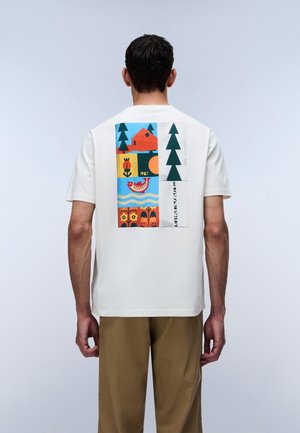 Mand iført hvid t-shirt med farverigt motiv af hus, træer, fisk, blomster og koordinater på ryggen, stående mod en ensfarvet baggrund.