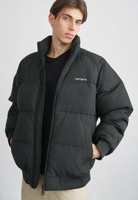 Fekete puffernadrág, magas gallérral, steppeltextúrával, cipzáras záródással és a Carhartt logójával a bal mellrészén. Bordázott mandzsetták és szegély.