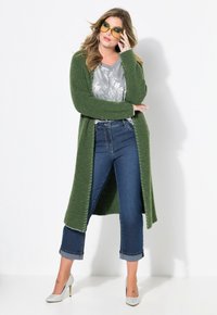 Lång grön stickad kappa, grå mönstrad topp, blå beskurna jeans med uppvikta fållar och silverfärgade högklackade skor. Gula solglasögon som en accent.