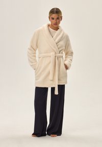 Jasno kremowy, teksturowany, pluszowy szlafrok z kołnierzem szalowym, z paskiem w talii i dwiema bocznymi kieszeniami, zestawiony z luźnymi ciemnymi spodniami.