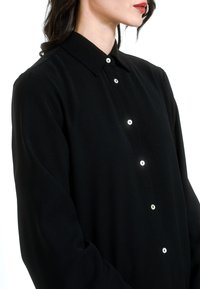 Black button-up shirt with a collar, made of smooth fabric. Features white buttons and long sleeves. No visible patterns or accents.