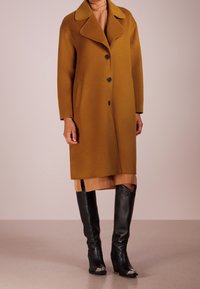 Mosterdkleurige wollen jas met brede revers, drie knopen en zijzakken, gedragen over een beige jurk en zwarte, kniehoge, gestructureerde laarzen.