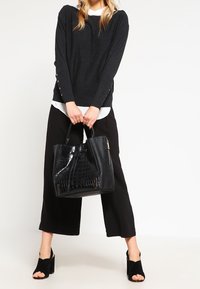 Borsa nera texturizzata con motivo coccodrillo e pelle liscia a contrasto, tenuta da una persona che indossa un maglione scuro e pantaloni neri larghi.