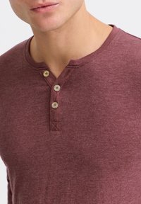 Maronfarvet langærmet Henley skjorte lavet af blødt, tekstureret stof med V-hals og placket med tre knapper. Har kontrasterende syninger.