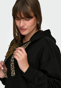 Juodas džemperis su kengūros kišene ir leopardų rašto raišteliais. Medžiaga yra minkšta, su laisvu kirpimu ir raišteliais rankogaliuose.