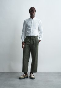 White button-up shirt, olive green pants, brown cap, and chunky shoes with cream accents. Smooth fabric, relaxed fit, and folded cuffs.
