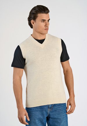 Beiger, ärmelloser Strickpullover mit V-Ausschnitt, kombiniert mit einem schwarzen T-Shirt darunter. Das Modell trägt blaue Jeans und präsentiert einen lässigen Look.