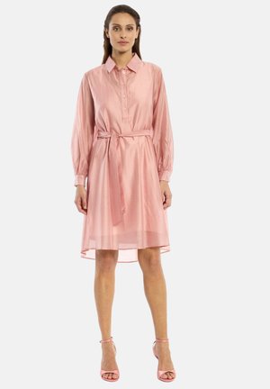 Vrouw draagt een lichtroze jurk tot op de knie met lange mouwen, een kraag, knopen aan de voorkant, een ceintuur in de taille en bijpassende roze sandalen met hoge hakken.