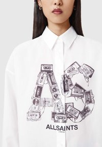 Valkoinen napillinen paita, jossa on graafinen printti pinotuista kasettinauhoista ja musta logo, jossa lukee "ALLSAINTS LONDON" alareunassa.