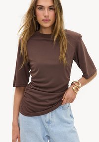 Jeune femme aux longs cheveux blonds portant un haut marron plissé à manches courtes, un jean bleu clair, ainsi que des bracelets et des boucles d'oreilles en or.