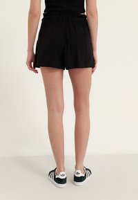 Personne portant un short noir à taille élastique et des baskets Adidas noir et blanc, debout dos à la caméra devant un fond uni.